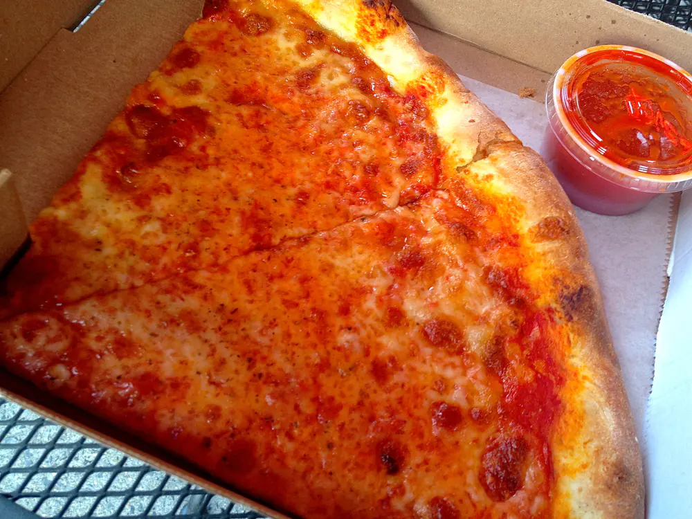 Two large triangular cheese slices of New York-style pizza in an open cardboard box beside a cup of tomato sauce