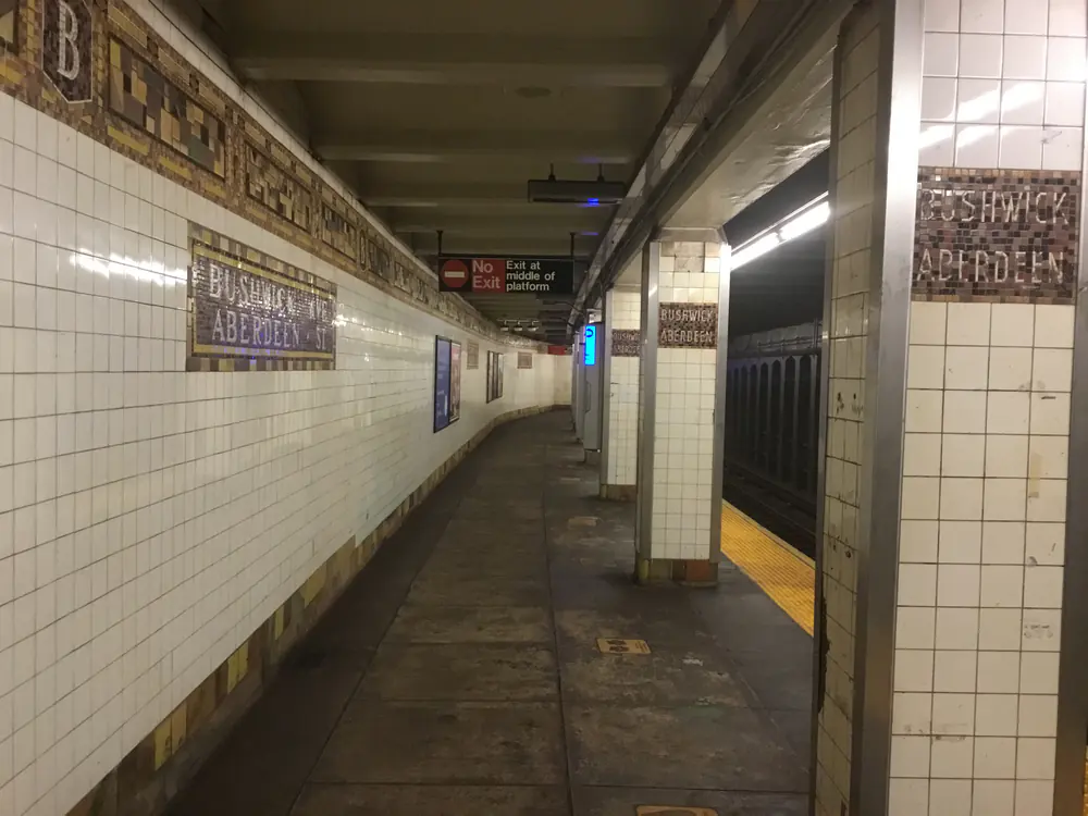 The tiled underground platform at Bushwick Avenue–Aberdeen Street station, with mosaic name tablets set into white subway tile