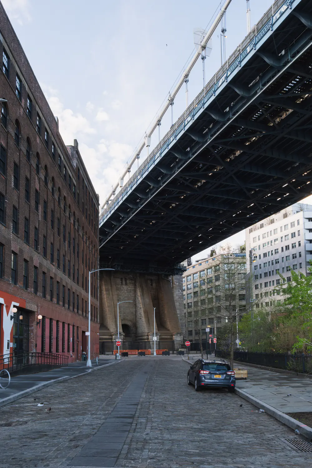 A cobbled DUMBO street running beneath the steel span of the Manhattan Bridge, framed by brick warehouse buildings