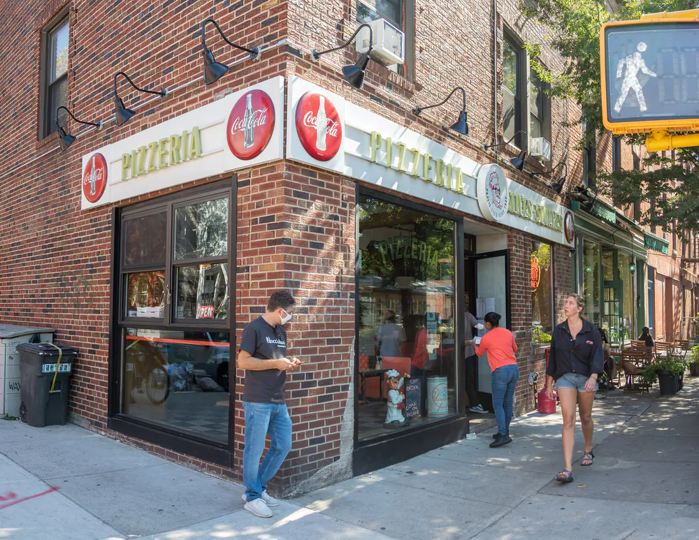 A brick corner pizzeria in Greenpoint with PIZZERIA signage and vintage Coca-Cola discs above the windows