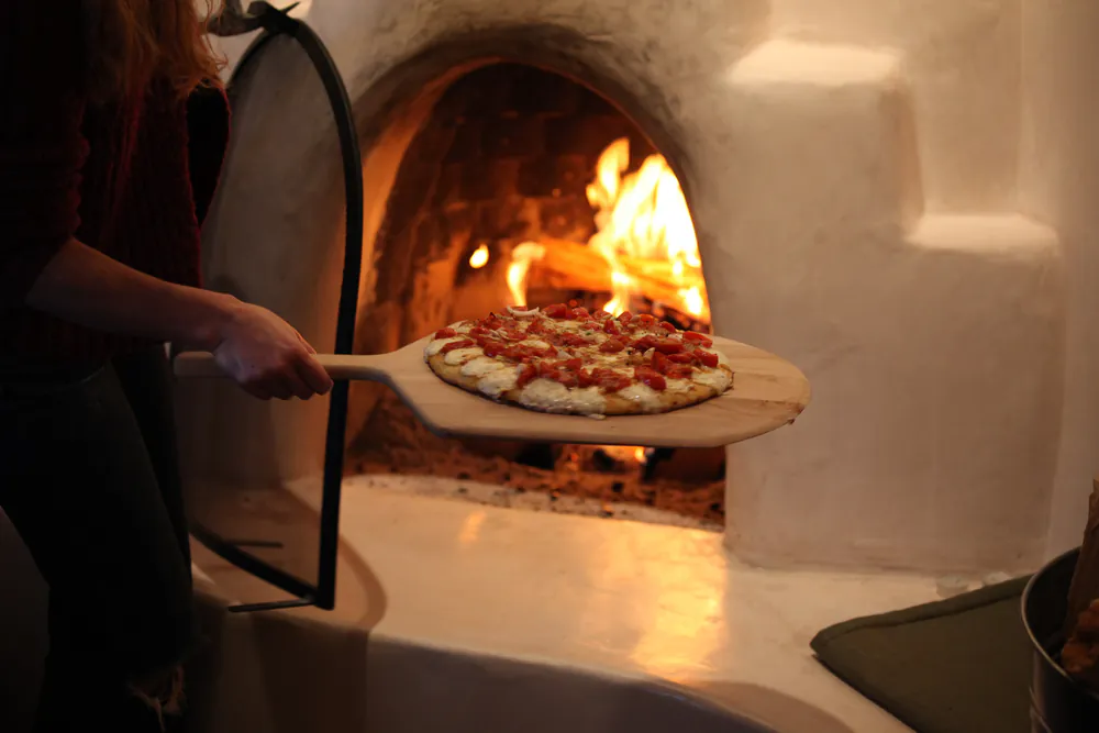 A pizza baking on the hearth floor of a wood-fired oven beside a bed of burning logs, with flame running along the low dome