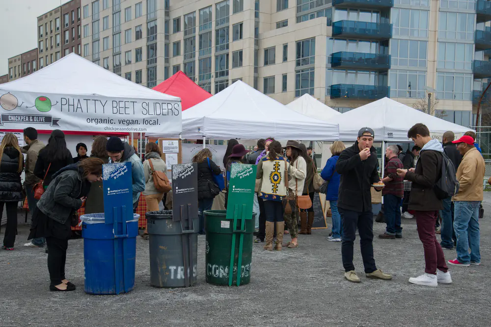 White vendor tents at an outdoor Brooklyn food market with a queue of customers and waterfront apartment blocks behind