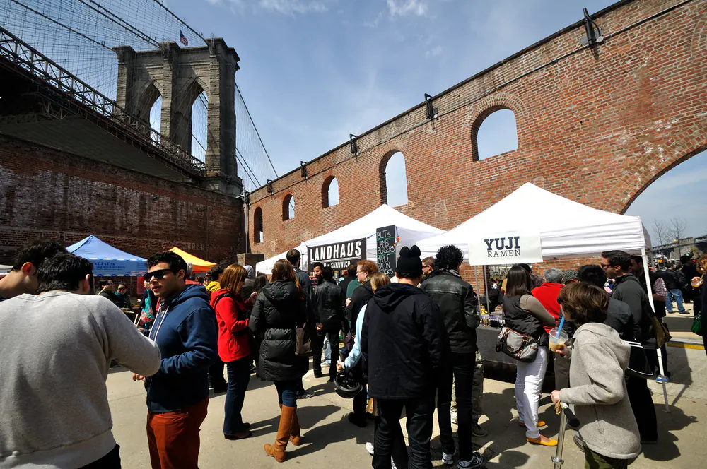 A crowd at white food-vendor tents beside brick warehouse arches in DUMBO, under the Brooklyn Bridge