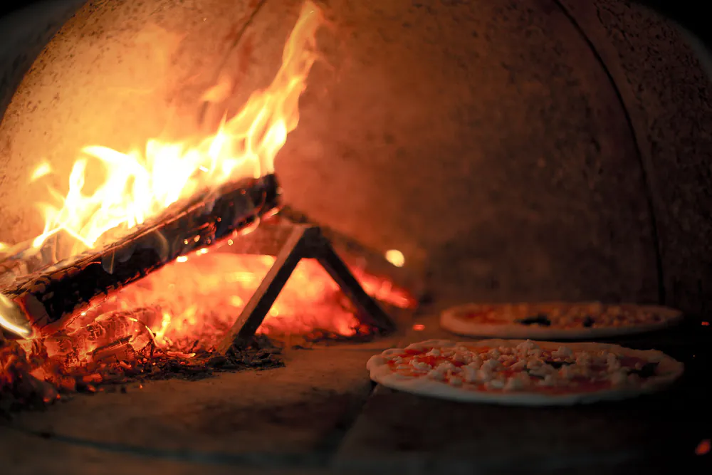 Flames curling across the ceiling of a wood-fired brick oven with a fire burning against the wall